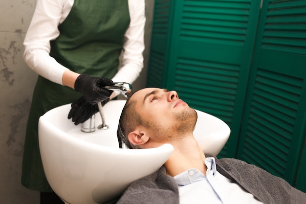 Il parrucchiere lava i capelli di un cliente prima di tagliarli. Uomo bello serio si lava la testa in un salone di bellezza