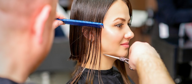 Il parrucchiere maschio taglia i capelli bagnati della giovane donna caucasica che pettina con un pettine in un parrucchiere