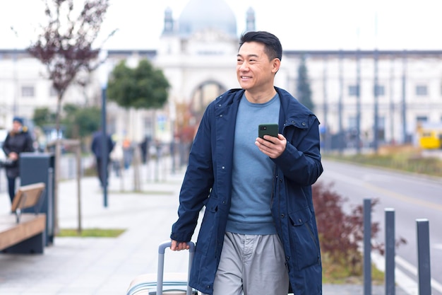 Il passeggero maschio turistico asiatico arriva dall'aeroporto cammina per la città con una valigia un uomo sorride e si rallegra usa il telefono e l'applicazione per prenotare un alloggio e prenotare un taxi