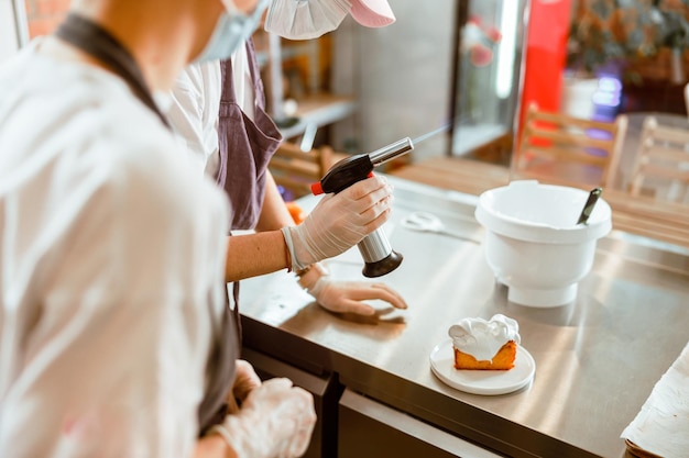 Il pasticcere con bruciatore a gas e il collega in maschera lavorano con il pan di spagna in panetteria