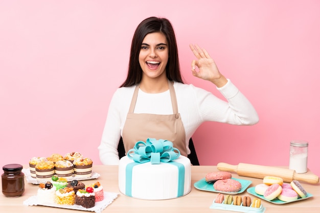 Il pasticcere con una grande torta in una tabella ha sorpreso e mostrando il segno giusto