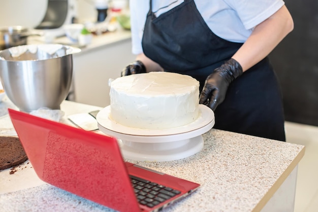 Il pasticcere cuoco pasticcere o fornaio in guanti neri e grembiule da cucina nero fa una torta. Torta di compleanno fatta in casa. Concetto di pasticceria fatta in casa, cucinare torte, hobby. Piccola impresa domestica femminile.