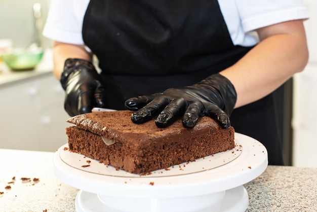 Il pasticcere cuoco pasticcere o fornaio in guanti neri e grembiule da cucina nero fa una torta. Torta di compleanno fatta in casa. Concetto di pasticceria fatta in casa, cucinare torte, hobby. Piccola impresa domestica femminile.