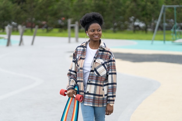 Il pattinatore della giovane ragazza africana tiene il longboard nella femmina urbana dello skate park in abiti casual nello skatepark