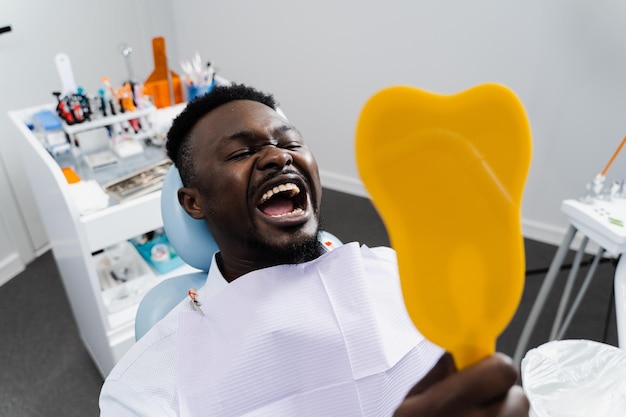 Il paziente africano si guarda allo specchio dopo aver rimosso la carie e riempito i denti Consultazione con il dentista nella clinica odontoiatrica Il paziente afroamericano sta visitando il dentista