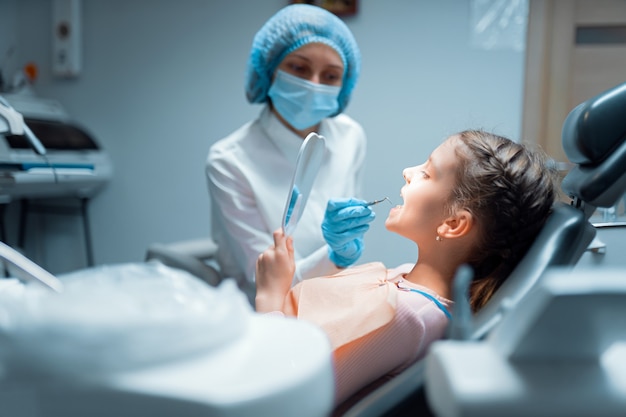 Il paziente della bambina e il dentista femminile stanno controllando i denti allo specchio dopo il trattamento in clinica odontoiatrica