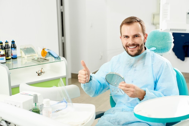 Il paziente soddisfatto con un sorriso bianco come la neve in una clinica dentale si siede su una sedia con uno specchio tra le mani