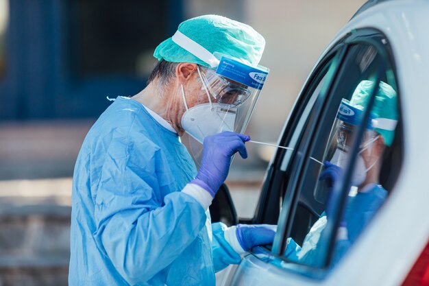 Il personale medico che indossa un DPI, esegue la PCR con un tampone in mano, su un paziente all'interno della sua auto per rilevare se è infetto da COVID-19