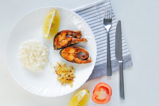 Il pesce fritto con riso alle cipolle e limone giace su un piatto su un asciugamano per cialde e posate su uno sfondo bianco...