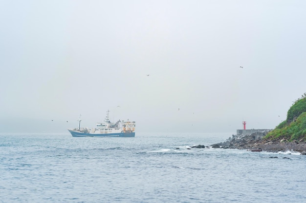 Il peschereccio emerge da dietro un promontorio con un faro che naviga in un mare nebbioso