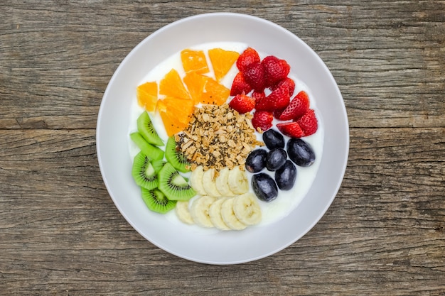 Il piatto di yogurt bianco naturale con i muesli, l'arancia, la banana, il kiwi, le fragole e l'uva fruttifica su fondo di legno. Yogurt e frutta come ingredienti intorno al piatto. Vista dall'alto. Concetto sano.