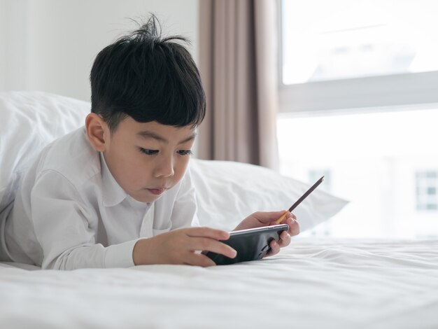 Il piccolo bambino asiatico sveglio ha messo a fuoco sullo smartphone mentre si trova sul letto