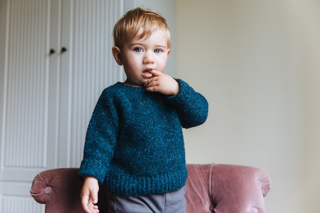 Il piccolo bambino biondo bello tiene il dito in bocca, guarda con i suoi occhi attraenti blu