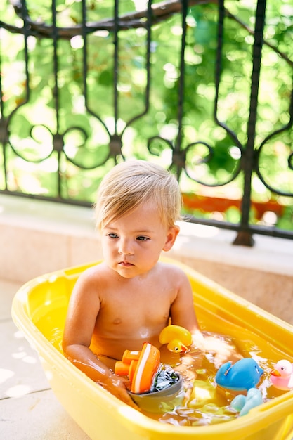 Il piccolo bambino con un mestolo in mano si siede in una bacinella con acqua e giocattoli