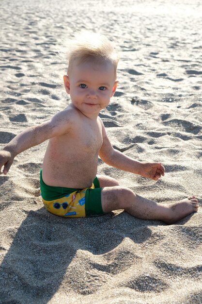 Il piccolo bambino si siede sulla soleggiata spiaggia sabbiosa e sorride