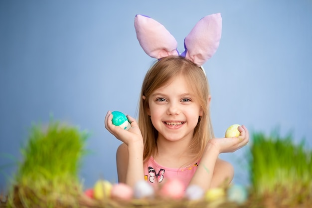 Il piccolo bambino sveglio indossa le orecchie del coniglietto il giorno di Pasqua. Bambina sveglia con le uova di Pasqua E l'erba verde. muro blu