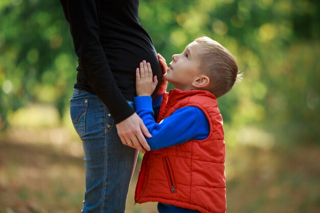 il piccolo figlio abbraccia la sua pancia incinta della mamma sulla natura. tocco felice del ragazzo una pancia della donna incinta all'aperto in parco verde. giovane famiglia felice in attesa del bambino. donna incinta che gode del tempo con il suo bambino