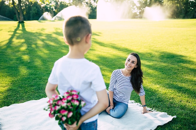 Il piccolo figlio carino fa una sorpresa con i fiori in mano per la sua giovane madre felice e bella all'aperto nel parco