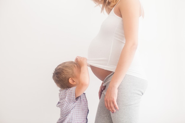 Il piccolo figlio guarda la pancia della madre incinta isolata su bianco. ciao piccola