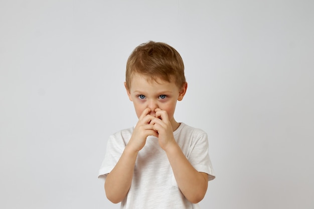 Il piccolo ragazzo in età prescolare si prende il naso con due dita su uno sfondo bianco