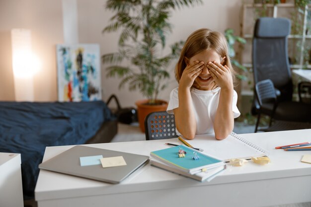 Il piccolo studente giocoso chiude gli occhi con i palmi delle mani alla scrivania con forniture e laptop a casa