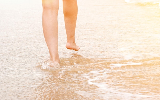 Il primo piano dei piedi femminili fa un passo sull'onda del mare e cammina sulla spiaggia sabbiosa Concetto di vacanze estive