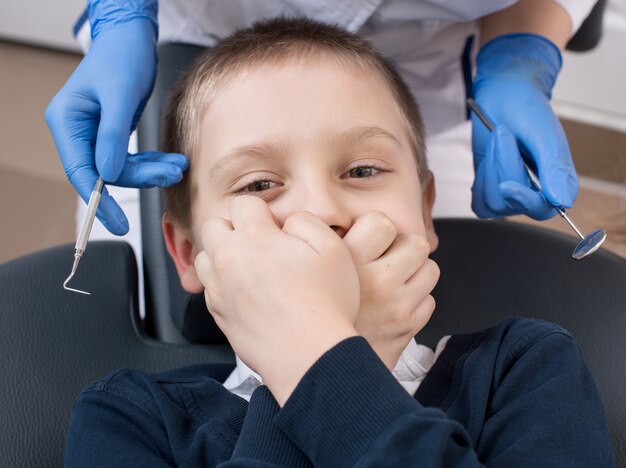 Il primo piano del ragazzo copre la sua bocca nella sedia del dentista e esamina la macchina fotografica