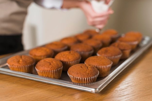 Il primo piano della donna che decora il fresco cuoce i muffin con crema sul vassoio