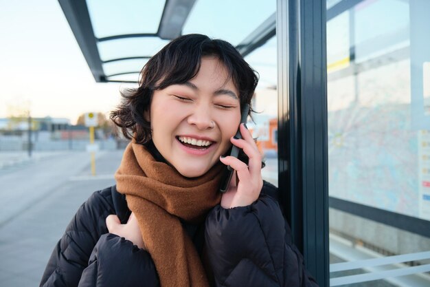 Il primo piano della ragazza asiatica alla fermata dell'autobus ride durante i colloqui di conversazione telefonica al telefono mentre attende
