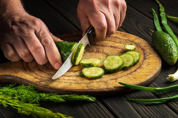 Il primo piano delle mani del cuoco con un coltello taglia un cetriolo fresco su un tagliere di legno
