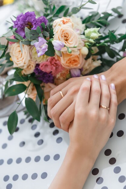 Il primo piano delle mani del matrimonio con anelli e bouquet giacciono sul tavolo