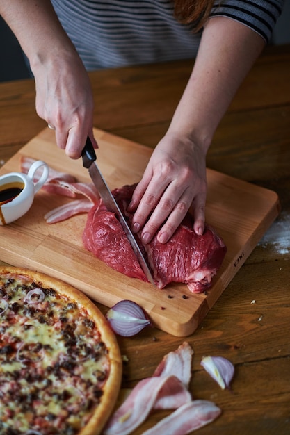 Il primo piano delle mani della donna ha tagliato il manzo con il coltello. Cipolla rossa, fette di prosciutto e pizza sul tavolo.