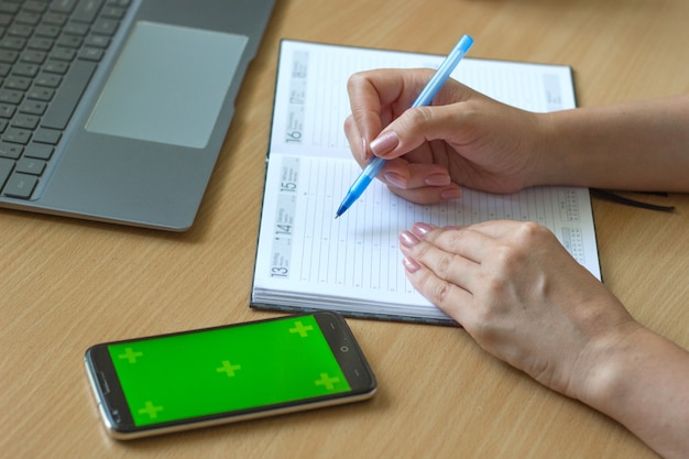 Il primo piano delle mani femminili scrive su un taccuino su un tavolo di legno uno smartphone e un laptop si trovano accanto ad esso