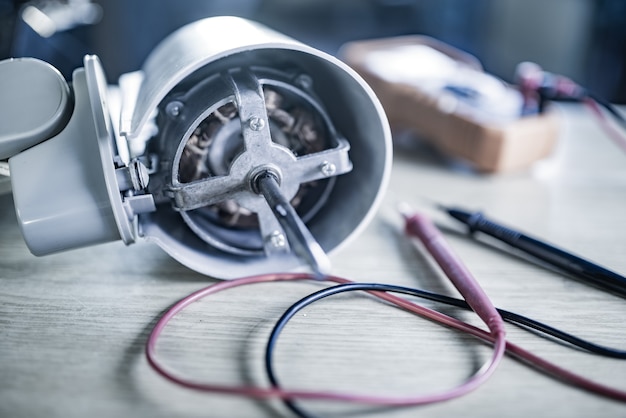 Il primo piano di un motore in ferro proveniente da una ventola di raffreddamento domestica e da strumenti di prova giace su un tavolo durante la riparazione in un'officina specializzata. Il concetto di riparazione e ripristino di apparecchiature danneggiate