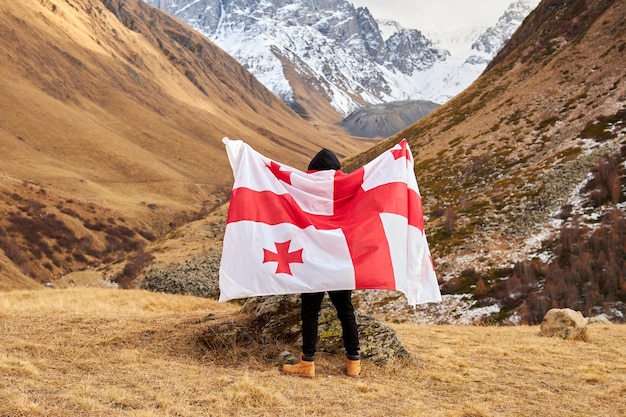 Il punto di vista posteriore del giovane felice che posa con la bandiera nazionale della georgia che sta all'aperto esamina il cappuccio della neve...