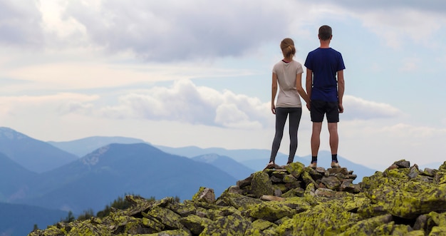 Il punto di vista posteriore di giovani coppie turistiche, l'uomo atletico e la ragazza esile stanno la mano della tenuta sulla cima della montagna rocciosa che gode del panorama strabiliante della montagna dell'estate. Concetto di turismo, viaggi e stile di vita sano.