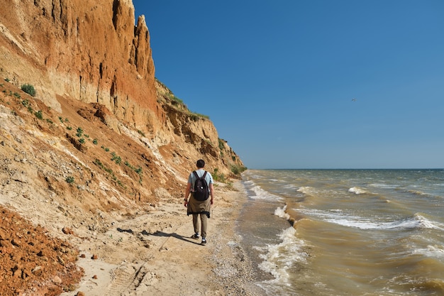 Il punto di vista posteriore di un giovane e di uno zaino cammina da solo lungo una collina di sabbia sopra il mare.