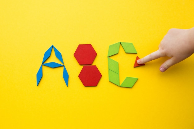 Il puzzle di legno del tangram di colore nelle lettere di alfabeto ABC modella su fondo giallo con la mano di un bambino