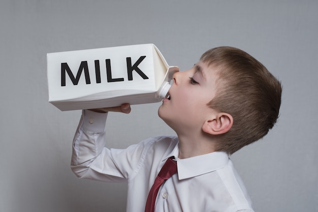Il ragazzino beve da un grande pacchetto di latte bianco. Camicia bianca e cravatta rossa.
