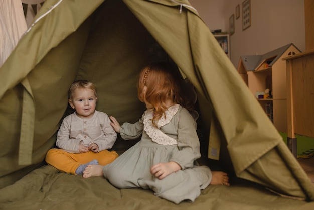 Il ragazzino biondo dalla pelle chiara sta guardando la fotocamera mentre è seduto in tenda con sua sorella dai capelli rossi in abito Concetto di infanzia