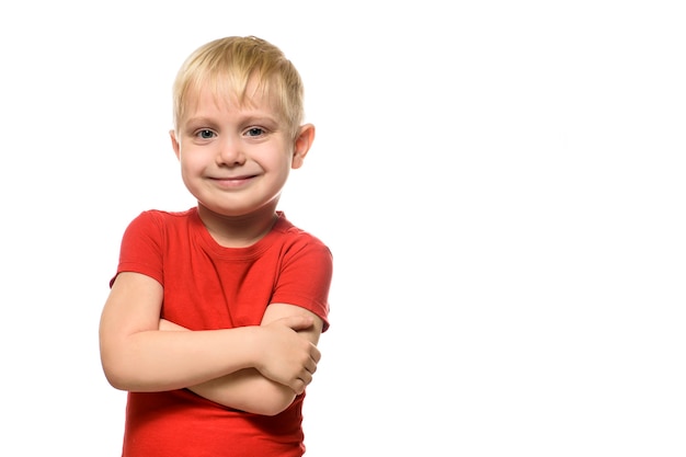 Il ragazzino biondo sorridente in una maglietta rossa sta con le braccia piegate. Isolare su sfondo bianco.