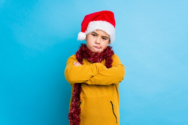 Il ragazzino che celebra il giorno di Natale che porta un fronte accigliato isolato cappello di Santa nel dispiacere, tiene le armi piegate.