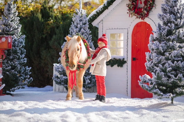 Il ragazzino con gli occhiali accarezza il pony adorabile vicino alla piccola casa di legno e agli alberi innevati.