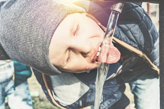 Il ragazzino divertente in cappello lavorato a maglia grigio beve l'acqua dalla pompa dell'acqua dell'annata nel cortile della casa del villaggio