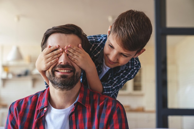 Il ragazzino felice in forma giocosa chiude gli occhi di suo padre mentre è seduto sul divano a casa.