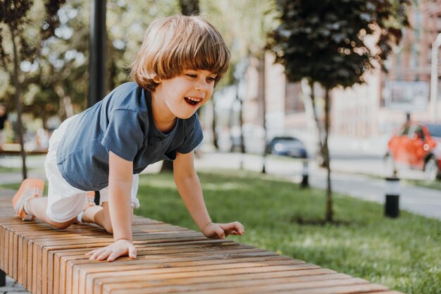 Il ragazzino grazioso del brunette in una maglietta blu e pantaloncini bianchi sta giocando sulla panca di legno nel parco. bambino che cammina all'aperto. concetto di salute. spazio per il testo. Foto di alta qualità