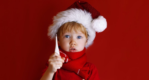 Il ragazzino in un cappello rosso della Santa tiene un termometro. Il bambino è malato prima di Natale