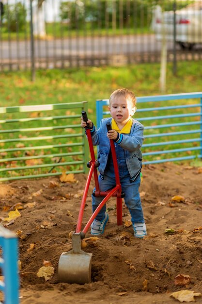 Il ragazzino in vestiti del denim gioca con un escavatore del giocattolo nel parco giochi nel parco d'autunno.