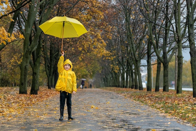 Il ragazzino solitario sta alzando il suo ombrello giallo nel parco autunnale piovoso Bambino su una strada piovosa con foglie che cadono