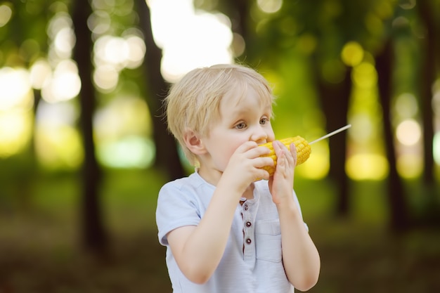 Il ragazzino sta mangiando mais all&#39;aperto. Cibo di strada per bambini.